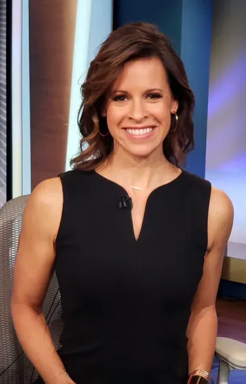 Jenna Wolfe with a friendly and engaging expression, wearing a sleeveless black dress. She has short, wavy brown hair and is accessorized with small hoop earrings and a delicate bracelet.