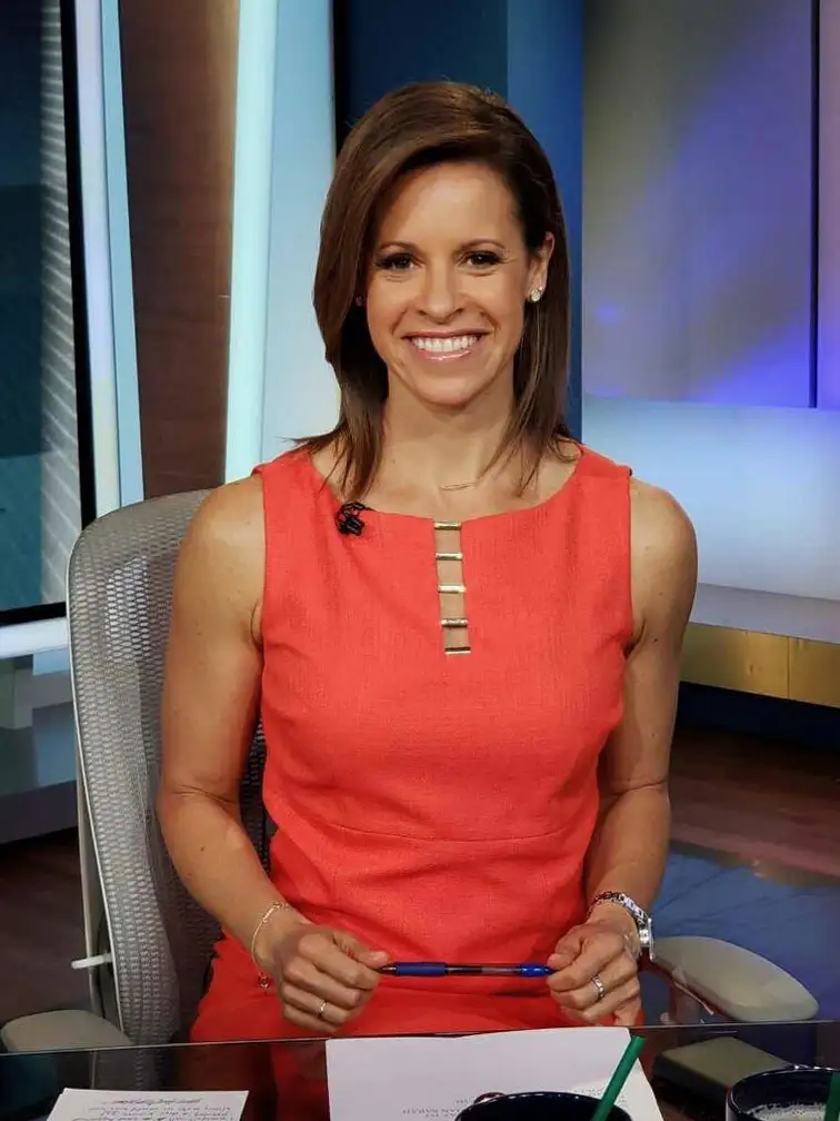 Jenna Wolfe sitting in a studio with a backdrop that reads “FIRST THINGS FIRST,” on a television show. She has a natural, warm expression and is positioned with her hands together on her lap. The setting has a modern, professional look typical of a TV studio.