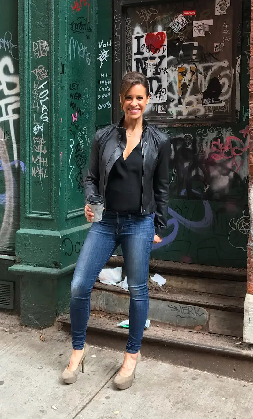 Jenna Wolfe in front of a doorway heavily adorned with graffiti and stickers, capturing a typical urban street art scene. She is smiling broadly, dressed in a casual chic style featuring a black leather jacket, a simple black top, and fitted blue jeans. She accessorizes her look with beige high-heeled shoes and is holding a coffee cup in one hand. Her outfit and the setting create a contrast that highlights both her stylish appearance and the vibrant, edgy background.